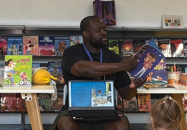Semi-close up photo from behind a group of kids watching illustrator Dapo Adeola showing books to them and the audience.