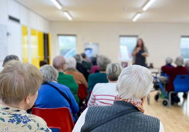 Portrait photo from back of room showing seated audience watching an author speak