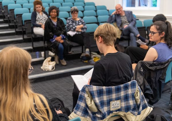 Photo of people, some from behind with a book open on lap and other people in background facing them as an audience