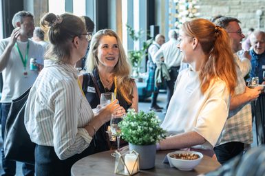 Three people chatting with drinks in hand in a hospitality space with plant on table in foreground and more people in background