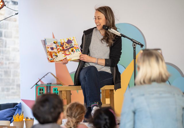 Photo of person holding up a picture book and reading with microphone on stand