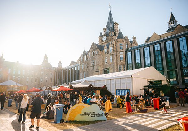 The Book Festival courtyard in the sun with crowds of people and sign that says 'Welcome to the Edinburgh International Book Festival'