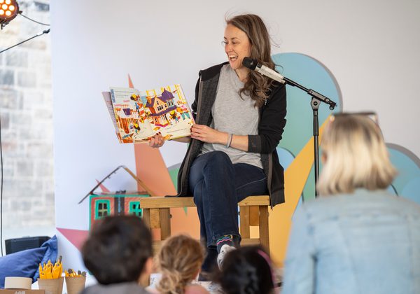 Speaker on stage holding up children's picture book as they read with backs of audience children in foreground