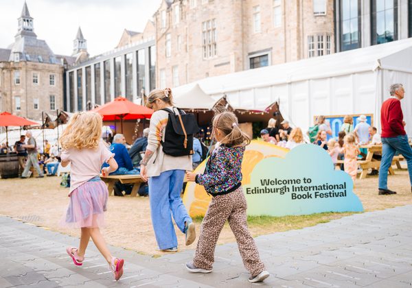 Adult and two kids walking into Book festival with sign that says 'Welcome to Edinburgh International Book festival' behind them