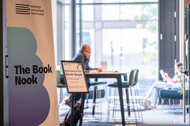 A sign that says 'the book nook' with one person sitting at a table and people in the background sitting reading