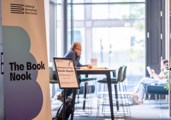 A sign that says 'the book nook' with one person sitting at a table and people in the background sitting reading